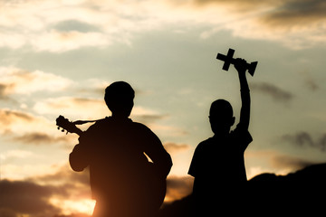 Two young christian worship God by holding cross and playing guitars with light sunset background,christian concept.