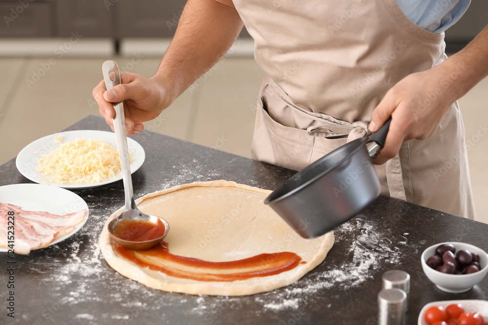 Young man applying sauce on pizza dough at table