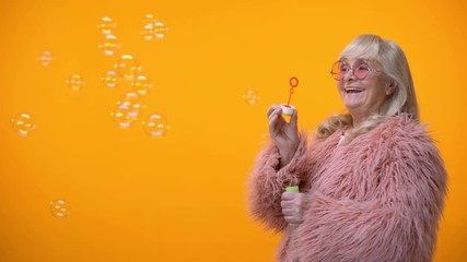 Sticker - Positive aged woman in funny pink coat and round sunglasses making soap bubbles