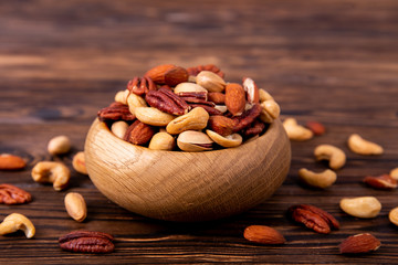 Wall Mural - Mixed nuts in wooden bowl and scattered on table. Trail mix of pecan, almond, macadamia & brazil edible nuts with walnut hazelnut on wood textured surface. Background, copy space, top view, close up.