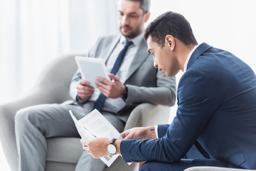 Wall Mural - young businessman holding document with business charts and colleague using digital tablet behind