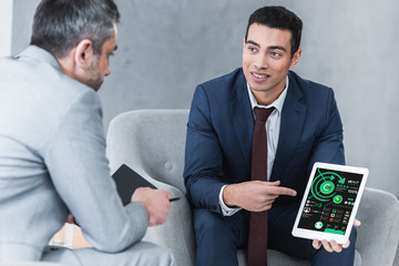 Wall Mural - smiling young businessman pointing at digital tablet with business charts on screen and looking at male colleague during conversation