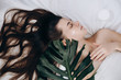 Attractive young long-haired girl applying cream on her face lying on the bed with a green leaf enjoying and relaxing  , day spa . Skin care and beauty concept.
