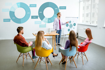 Young male speaker reporting to the audience during the meeting in the conference room with charts on the background