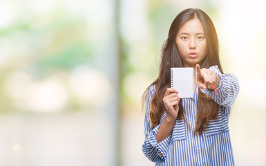 Sticker - Young asian woman holding notebook over isolated background pointing with finger to the camera and to you, hand sign, positive and confident gesture from the front