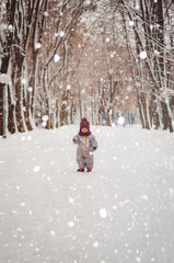 Canvas Print - Portrait of a cute baby dressed in a gray jacket and a red hat that walks through the snow covered snow park. She smiles one in the photo during the snowfall