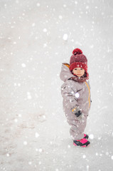 Wall Mural - Portrait of a cute baby dressed in a gray jacket and a red hat that walks through the snow covered snow park. She smiles one in the photo during the snowfall