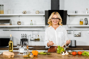 smiling attractive mature woman mixing salad in glass bowl in kitchen and looking at camera