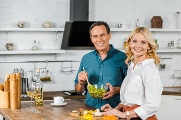 happy mature wife and husband cooking salad together in kitchen, looking at camera