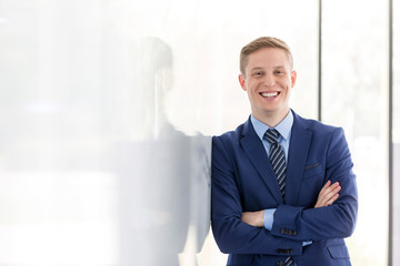 Portrait of smiling young businessman with arms crossed leaning on wall at office