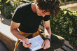 © iuricazac - Top view of handsome man student with curly hair, wearing black t-shirt sitting on the city street and preparing for exams. Freelancer businessman planning the day on the paper.