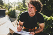 © iuricazac - Portrait of man with curly hair, writing some notices on paper and preparing for exams, wearing black t-shirt sitting on the city street. Freelancer businessman makes plans for new projects.