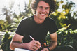 © iuricazac - Happy smiling young man with curly hair, writing some notices on paper and preparing for exams, wearing black t-shirt sitting on the city street. Freelancer businessman makes plans for new projects.