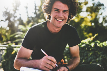 Wall Mural - Happy smiling young man with curly hair, writing some notices on paper and preparing for exams, wearing black t-shirt sitting on the city street. Freelancer businessman makes plans for new projects.