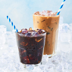 Summer drink iced coffee in a glass and ice coffee with cream in a tall glass surrounded by ice on white marble table over blue background. Selective focus, copy space for text. Square crop.