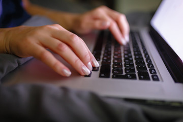 Close-up of woman's hand using laptop with blank screen on bed in home interior. The light from the screen illuminates the female hands on the laptop keyboard. Freelancer works at home in the evening