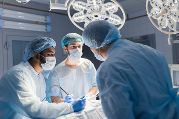 Canvas Print - Male surgeon holding instrument in abdomen of patient. Female and male assistants help him. Indian surgeon performing laparoscopic surgery in operating room. Minimally invasive surgery. Blue filter