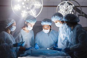 Wall Mural - Surgeons team during operation process in surgery in a dark background. Surgeons stand up through 10-,12-,15-hour surgeries without food, without bathroom breaks without complaints.