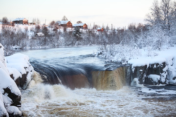 Wall Mural - View of a flat waterfall with village on the hill. Sunset at winter season. District of the village Nadvoitsy, Segezha, Karelia, Russia