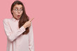 Scared emotive young woman with black hair, points aside, has fearful expression, demonstrates something exciting, models against pink background. People, advertisement or promotion concept.