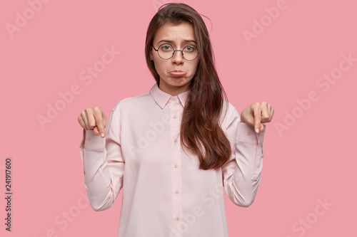Fotografia, Obraz Horizontal shot of displeased young woman has gloomy facial expression, points at floor with both index fingers, feels dislike, wears elegant clothes, isolated over pink background