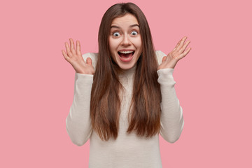 Wall Mural - Headshot of attractive excited woman spreads palms near face, has surprised expression, has long hair, dressed casually, isolated over pink studio wall. Happy Caucasian lady feels overjoyed, gestures