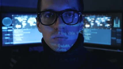 Portrait of Hacker programmer in glasses looks at camera while blue code characters reflect on his face in cyber security center filled with display screens.
