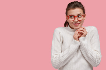 Wall Mural - Optimistic dreamy young woman keeps hands together, focused aside with gentle smile, has intention or plan for something grandiose, wears glasses for good vision, isolated over pink studio wall