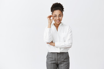 Wall Mural - Good-looking stylish and smart female psychologist with dark skin in white-collar shirt and pants, touching glasses and smiling with self-assured expression, gazing at camera