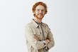 Studio shot fo confident friendly casual male redhead with beard and wavy hairstyle crossing hands on chest and smiling joyfully at camera gazing with satsfied and pleased look over gray wall