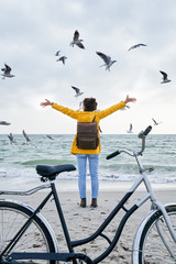 back view to a young female standing with a bicycle on the beach with raised hands surrounded with flying seagulls.