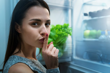 Girl getting secretly to eat from fridge at night, asking to keep silent. Unhealthy eating concept