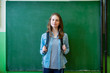 Young confident smiling female high school student standing in front of chalkboard in classroom, wearing backpack, looking at camera. Waist up portrait.