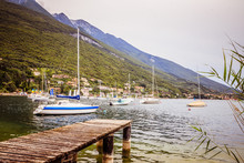 Wooden Dock Pier Extending Over Blue Lake Water, Sailing Boats And Mountains