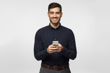 young business man standing isolated on gray background, holding smartphone, looking at camera and s