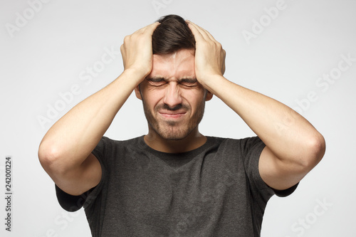 Cuadros en Lienzo Young man isolated on gray background, showing how much head hurts, experiencing pain, looking miserable and exhausted