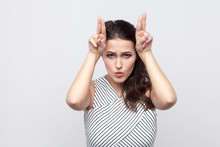 Portrait Of Funny Beautiful Young Brunette Woman With Makeup And Striped Dress Standing With Cow Gesture And Looking At Camera. Indoor Studio Shot, Isolated On Grey Background.