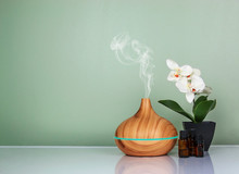 Electric Essential Oils Aroma Diffuser, Oil Bottles And Flowers On Light Green Surface With Reflection