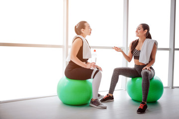 Wall Mural - Tired but happy young women sit at fitballs and talk. They smile to each other. Models have white towels on their shoulders. They sit in fitness room.