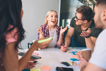 Cheerful young blonde woman smile and look forward. She wave with hands. Brunette woman and two young men look at her. They listen to. People work together in coworking.