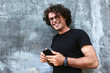 © Z A C - Happy smiling man standing outdoors texting on mobile phone. Young male with curly hair wears spectacles resting outside in the city browsing on his cell phone on concrete gray background.
