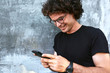 © iuricazac - Closeup image of smiling man standing outdoors texting on mobile phone. Young male with curly hair wears spectacles resting outside in the city browsing on his cell phone on concrete gray background.