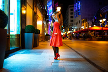 A Beautiful Mature Woman Standing In The Streets Of The City And Posing In A Red Dress.