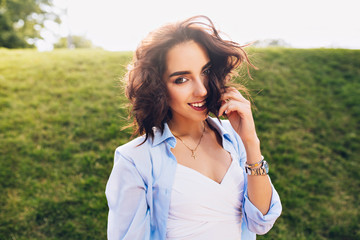 Portrait of cute brunette girl with short hair posing to camera in park on grass background. She wears white T-shirt and blue shirt. She is smiling.