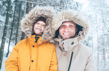 Wall Mural - Father and son dressed in Warm Hooded Casual Parka Jacket Outerwear walking in snowy forest cheerful smiling faces portrait. Father and son relatives and winter outfit concept image.