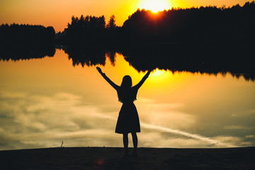 Wall Mural - Silhouette of a young beautiful girl with hands up against the background of the sunset in the reflection of the pond