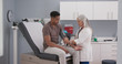 Mature female medical practitioner measuring blood pressure with monitor and stethoscope of a young male patient. Attractive black man having his blood pressure measured by senior white doctor