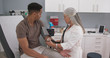 Senior caucasian doctor using stethoscope and gauge to measure patients blood pressure. Young black male seated in medical clinic having his blood pressure measured by mature female doctor