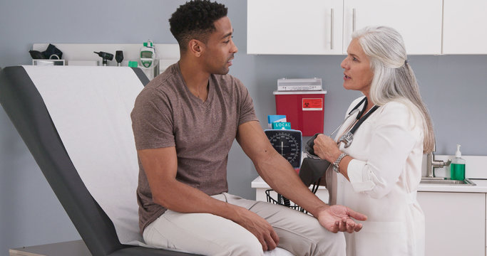 Wall Mural -  - Handsome african-american patient seated in medical room having his blood pressure checked by senior doctor. Mature white female physician checking male patients blood pressure in health clinic