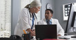 © Mark Adams - Portrait of millennial black doctor using computer while senior colleague directs him. Two doctors working inside medical office looking at computer monitor and typing